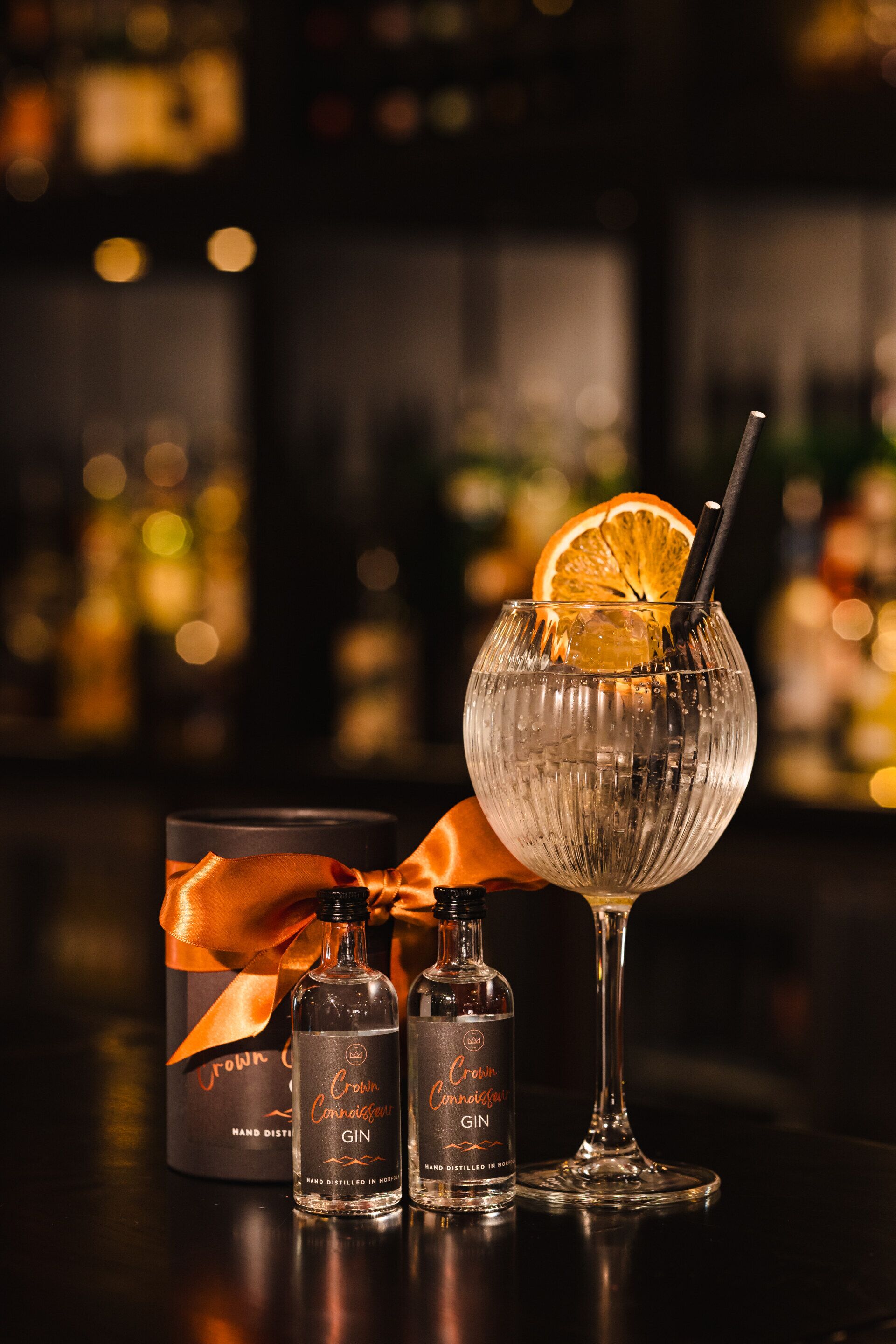 A bottle of Crown Connaught Gin, a bottle of Crown Connaught Gin, and a glass of gin with an orange slice and a straw.