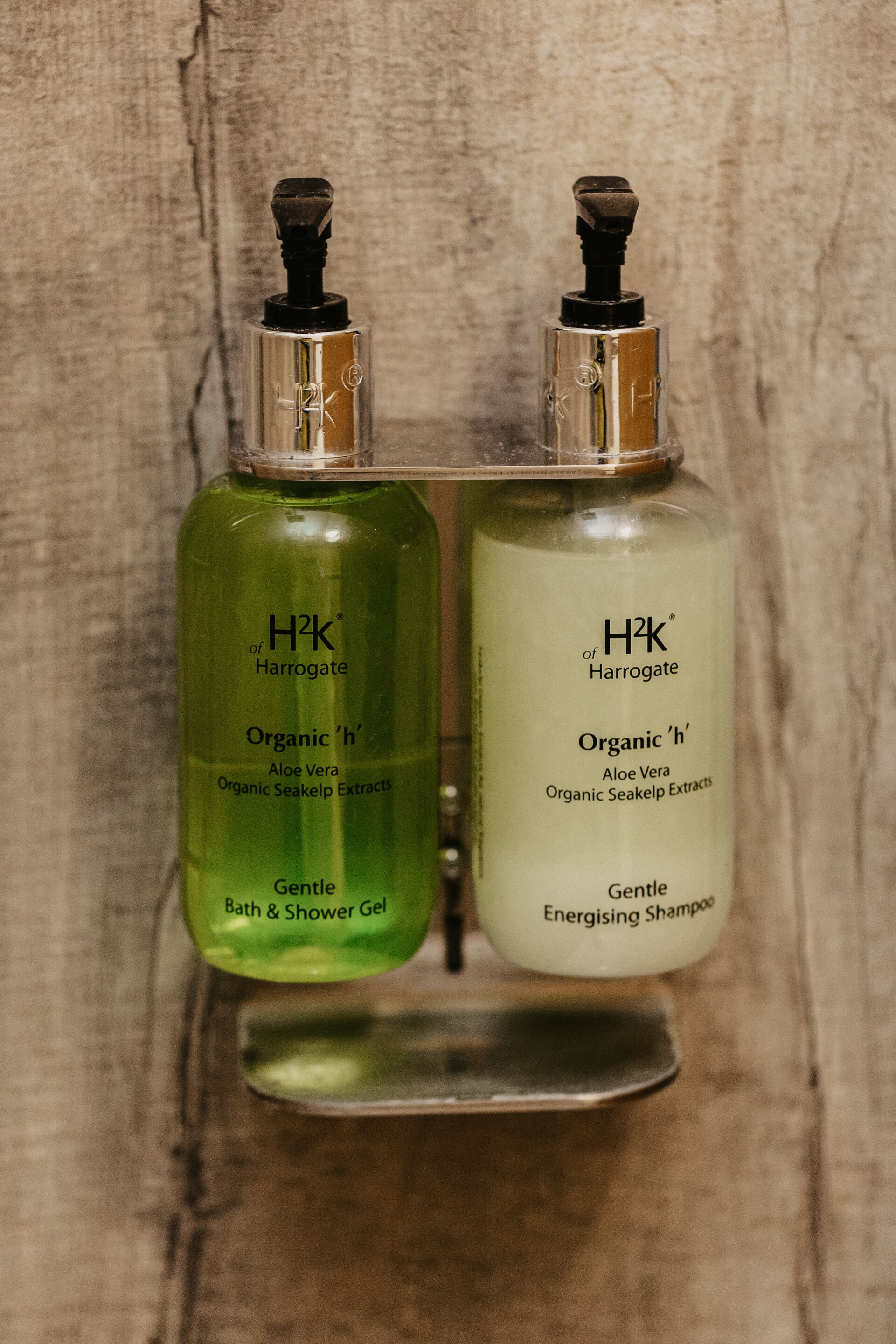 Two H2K of Harrogate bottles, one green and one white, labeled as Organic 'n' Aloe Vera Bath & Shower Gel and Organic 'n' Aloe Vera Energising Shampoo, respectively, on a wooden surface.