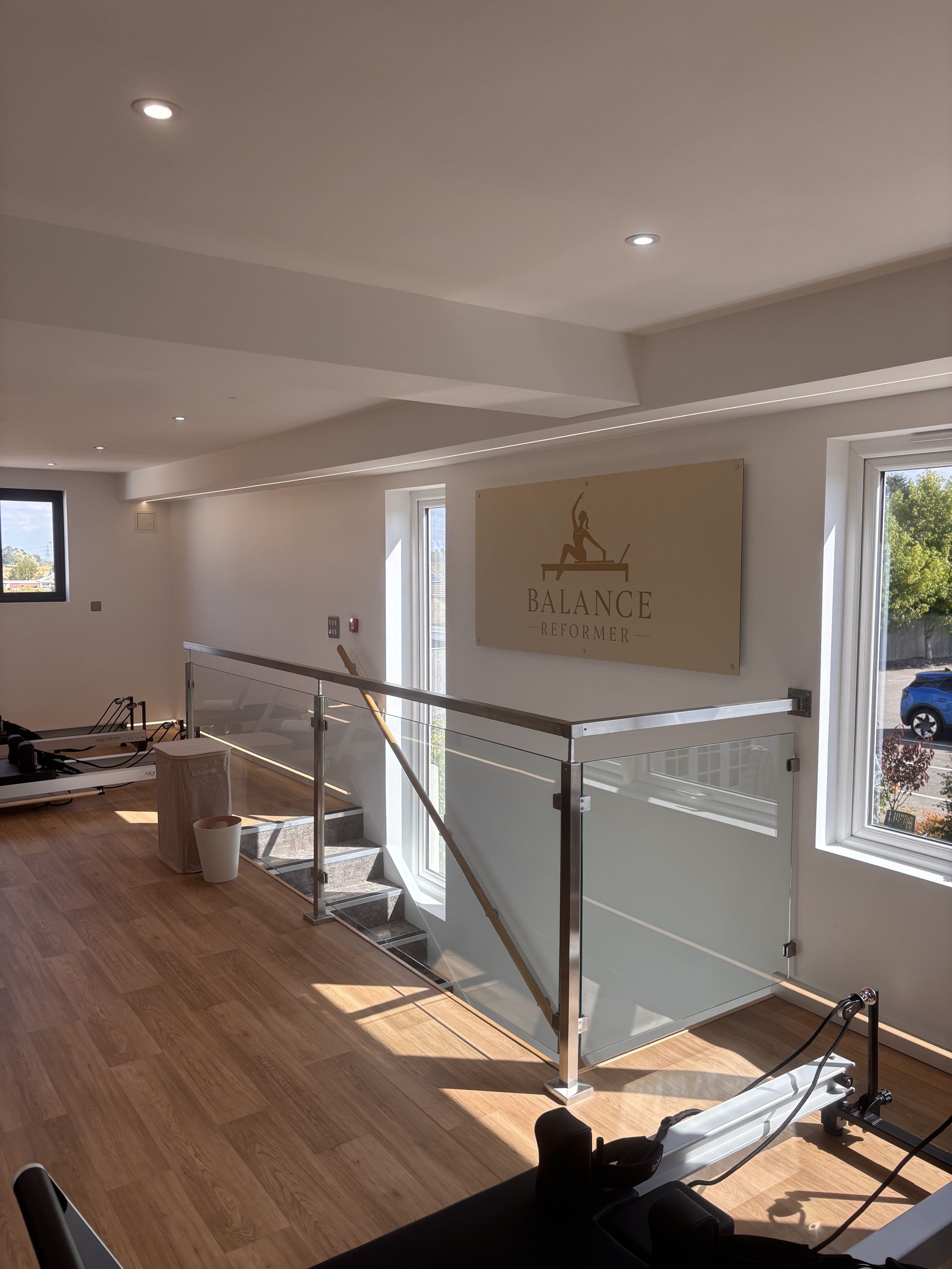 A modern interior with a wooden floor, a glass railing staircase, and a sign reading 'BALANCE - REFORMER'.