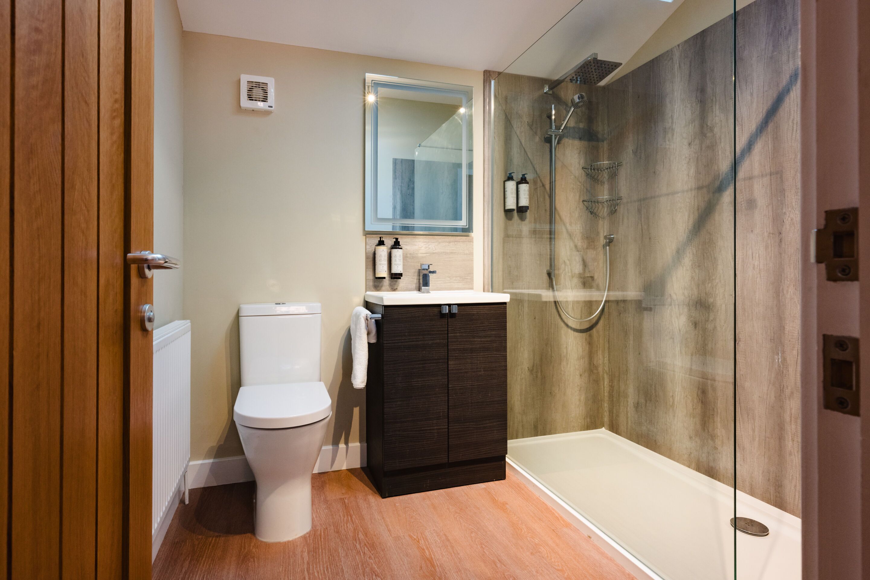 A modern bathroom with a toilet, dark wooden cabinet, glass shower enclosure, and a mirror above the sink.