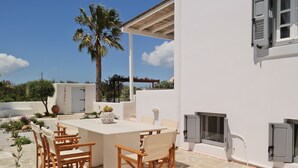 An outdoor patio with a white table and wooden chairs, surrounded by a white building with a stone floor.