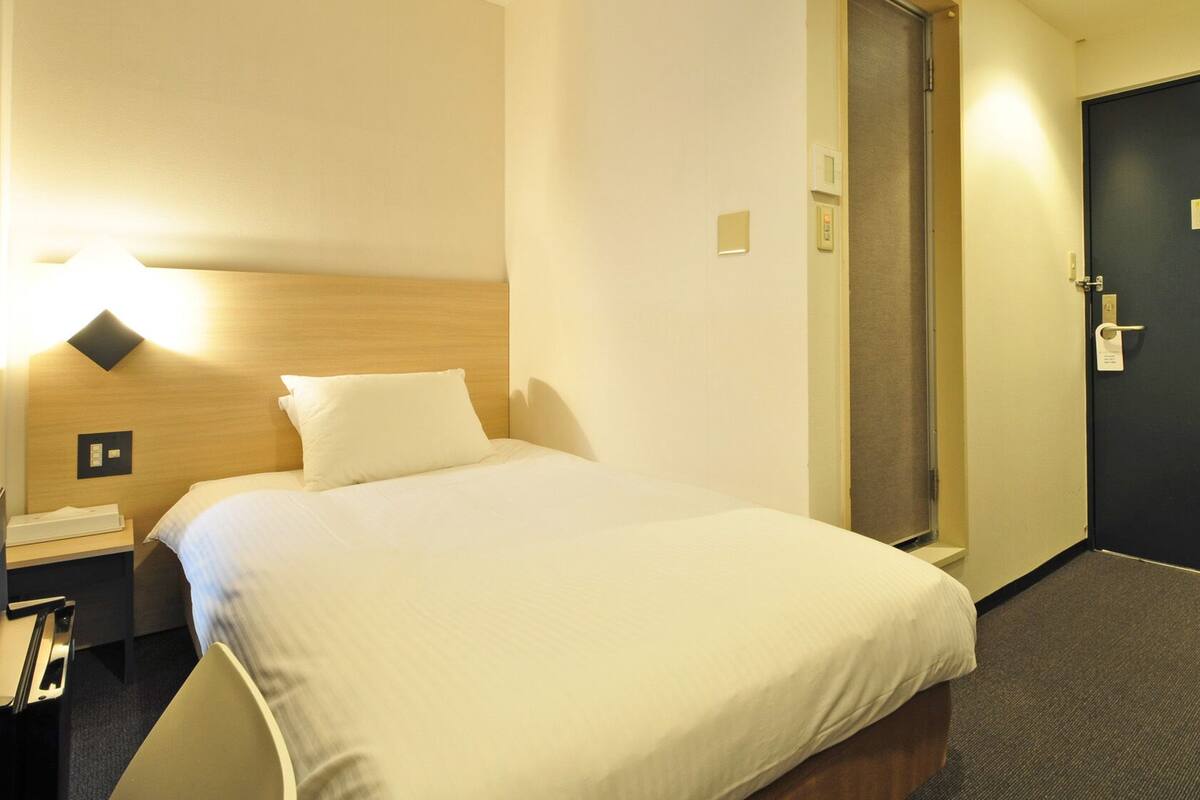 A hotel room with a bed, a desk, a chair, a door, and a wall-mounted light.