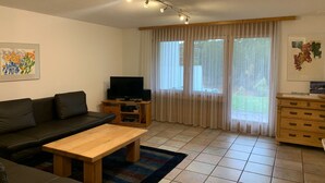 Ein Wohnzimmer mit einer Couch, einem Couchtisch, einem Fernseher auf einem Schrank, einer Holzeinrichtung und einem Fenster mit Vorhängen.