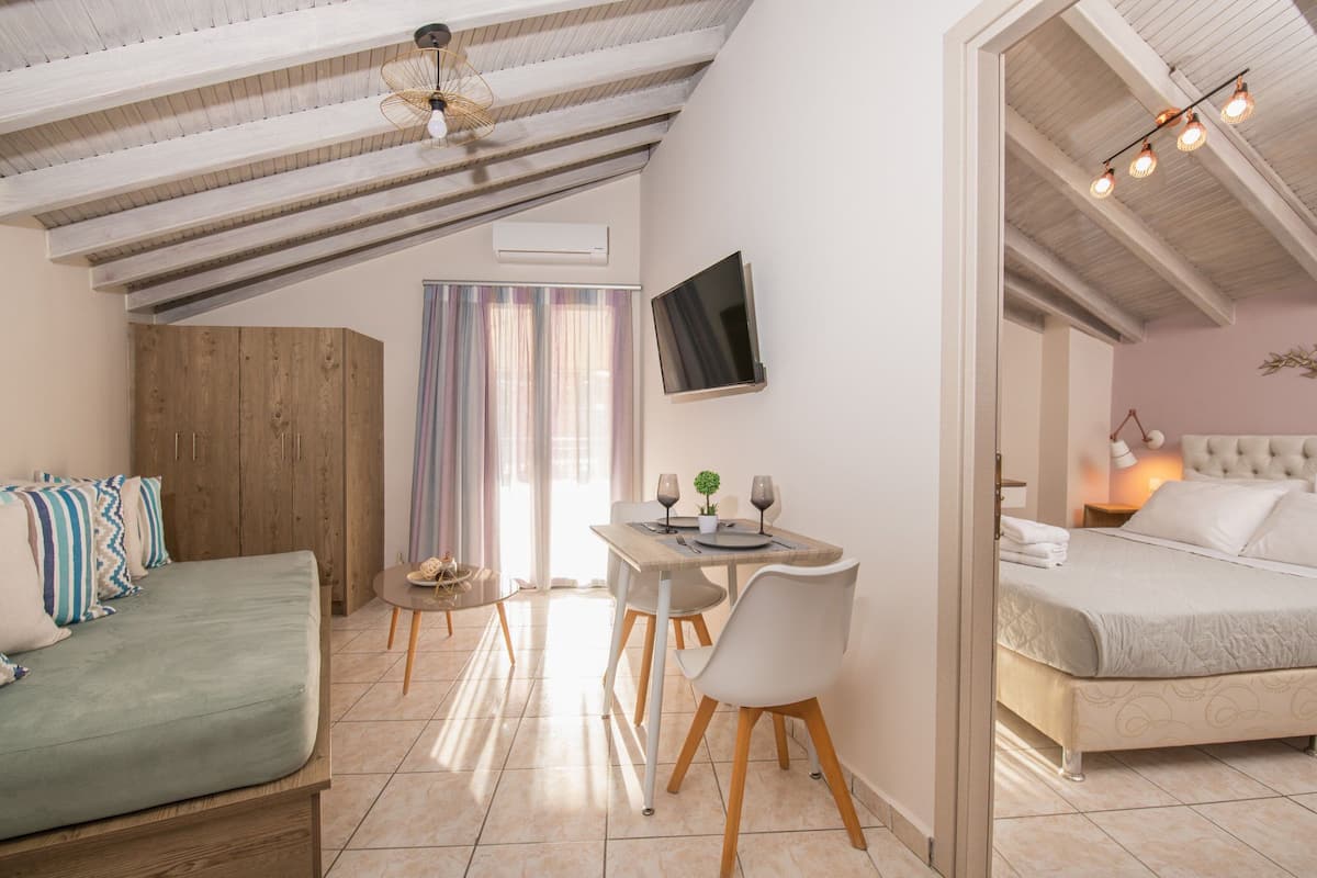 Une chambre confortable avec un lit, un petit coin salon avec une table et des chaises, un téléviseur fixé au mur et un plafond avec des poutres apparentes.