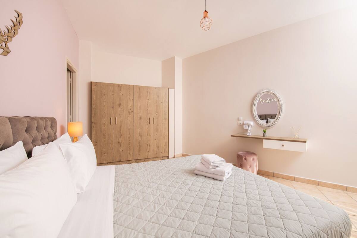 Une chambre avec un grand lit, une armoire en bois, une table de chevet avec une lampe, un miroir et un petit tabouret.