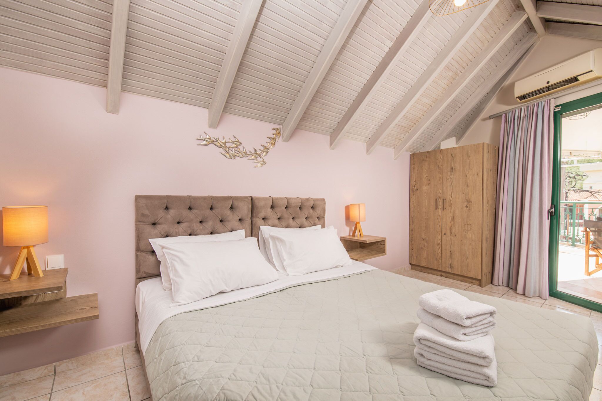 A bedroom with a large bed, wooden headboard, two bedside lamps, a wooden wardrobe, and a view through a glass door.