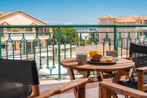 Een houten tafel met een bord eten, een kopje en een glas sinaasappelsap op een balkon met groene relingen.