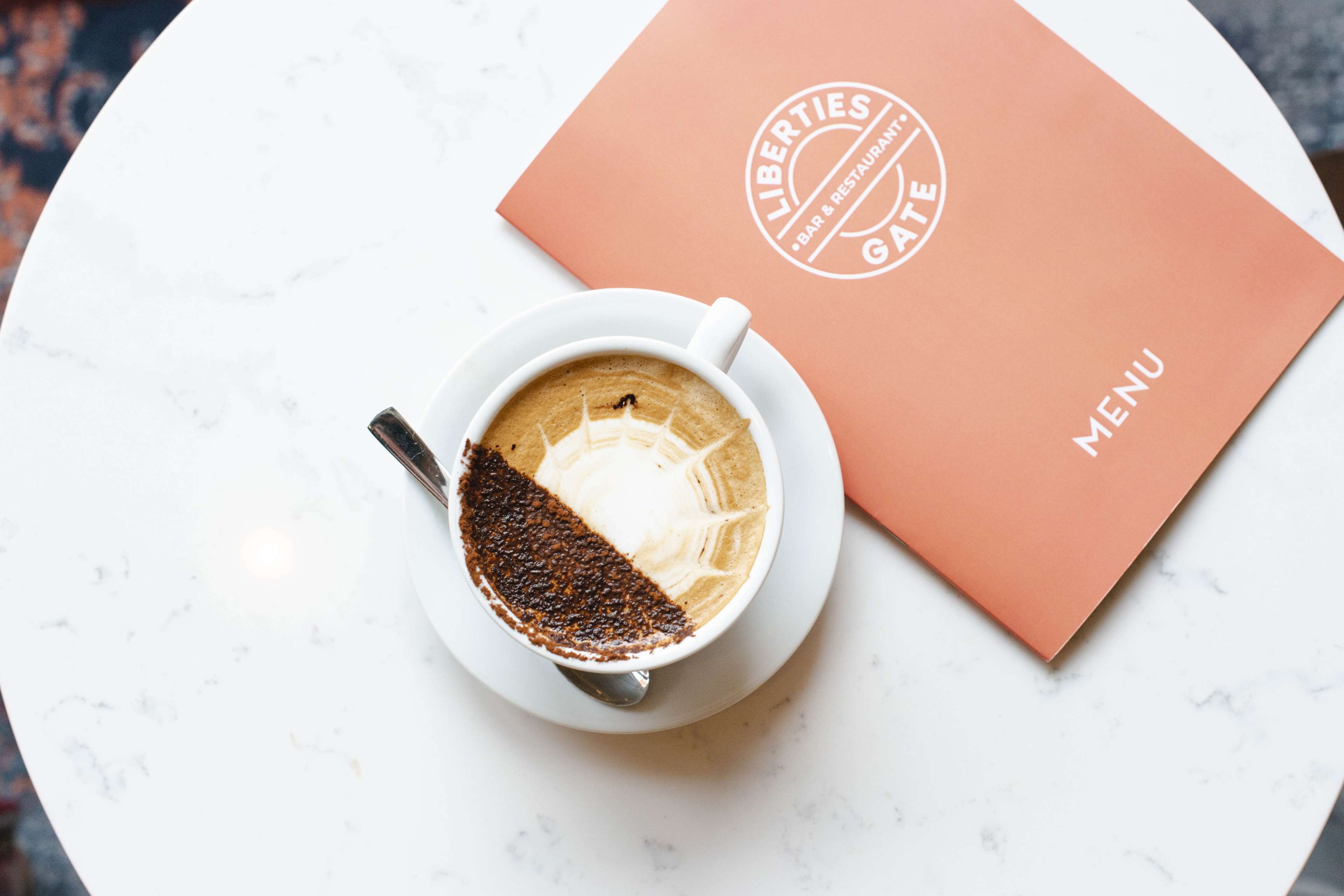 Eine Tasse Kaffee mit Latte-Art auf einem weißen Teller, platziert auf einem weißen Tisch neben einer Speisekarte mit dem Text 'LIBERTIES GATE BAR & RESTAURANT MENU'.