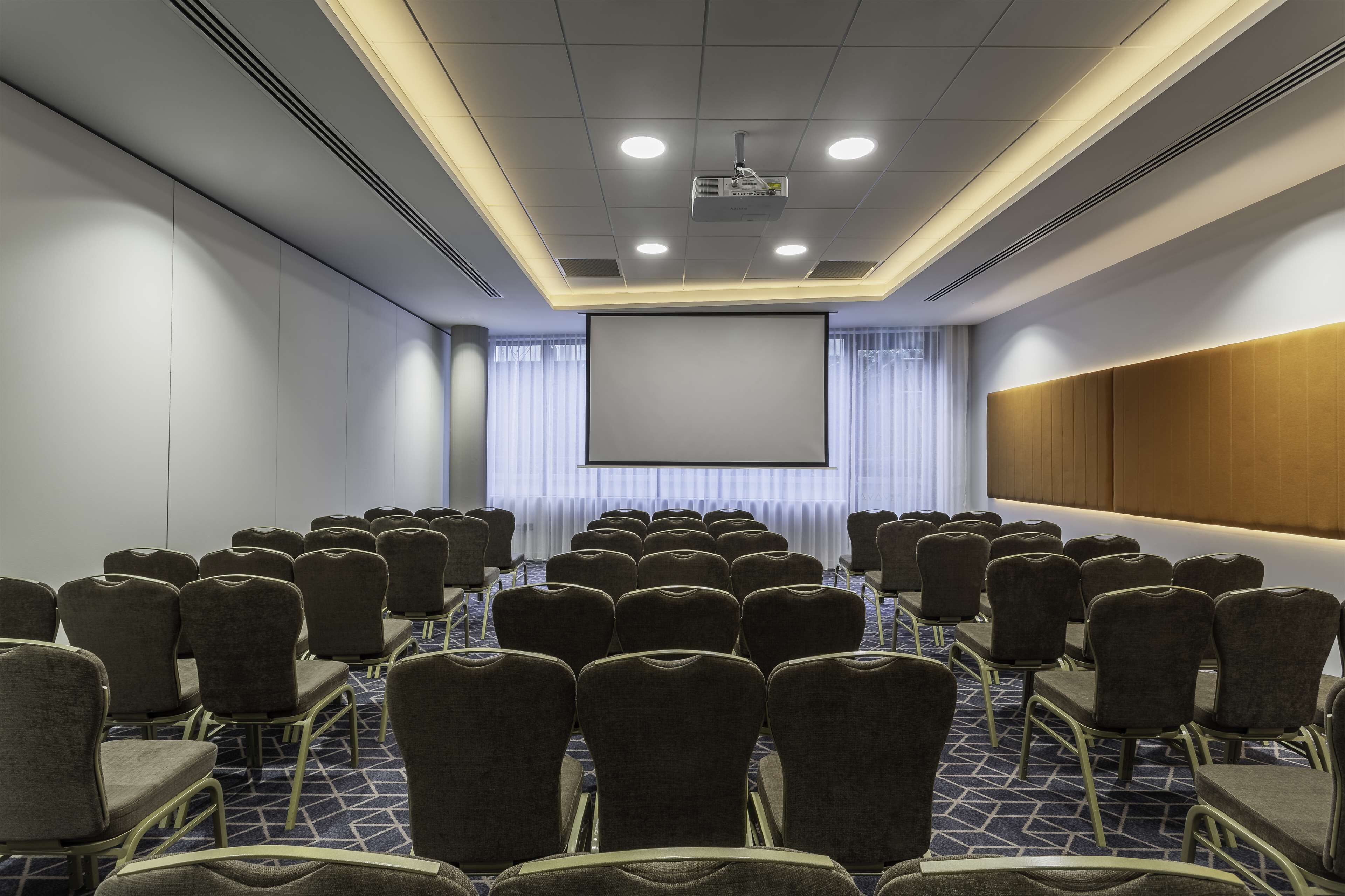 Sala de conferência com fileiras de cadeiras voltadas para uma tela grande, um projetor e uma parede revestida de madeira.