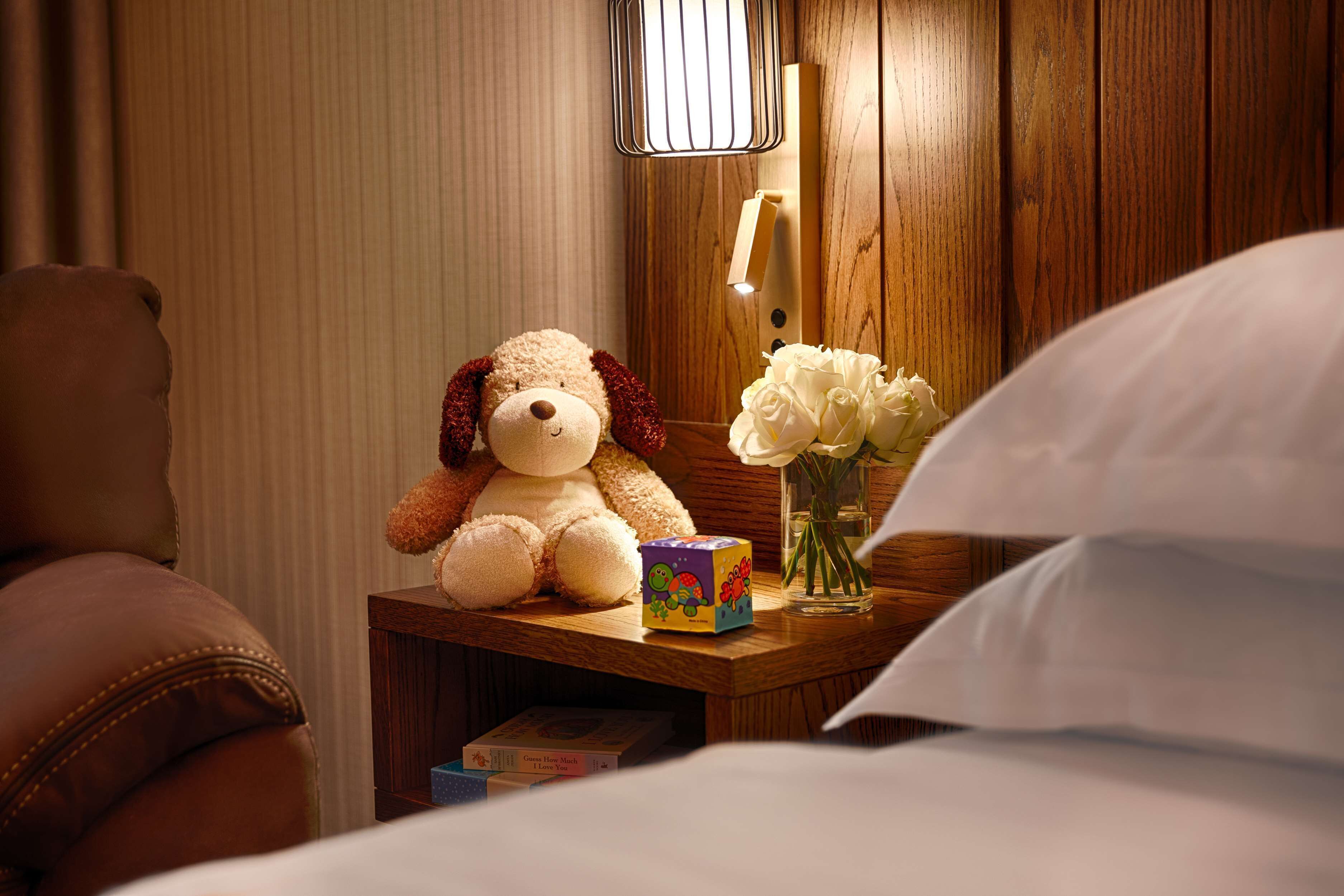 A neatly made bed with white sheets, a wooden bedside table with a teddy bear, a vase of white flowers, and a lamp.