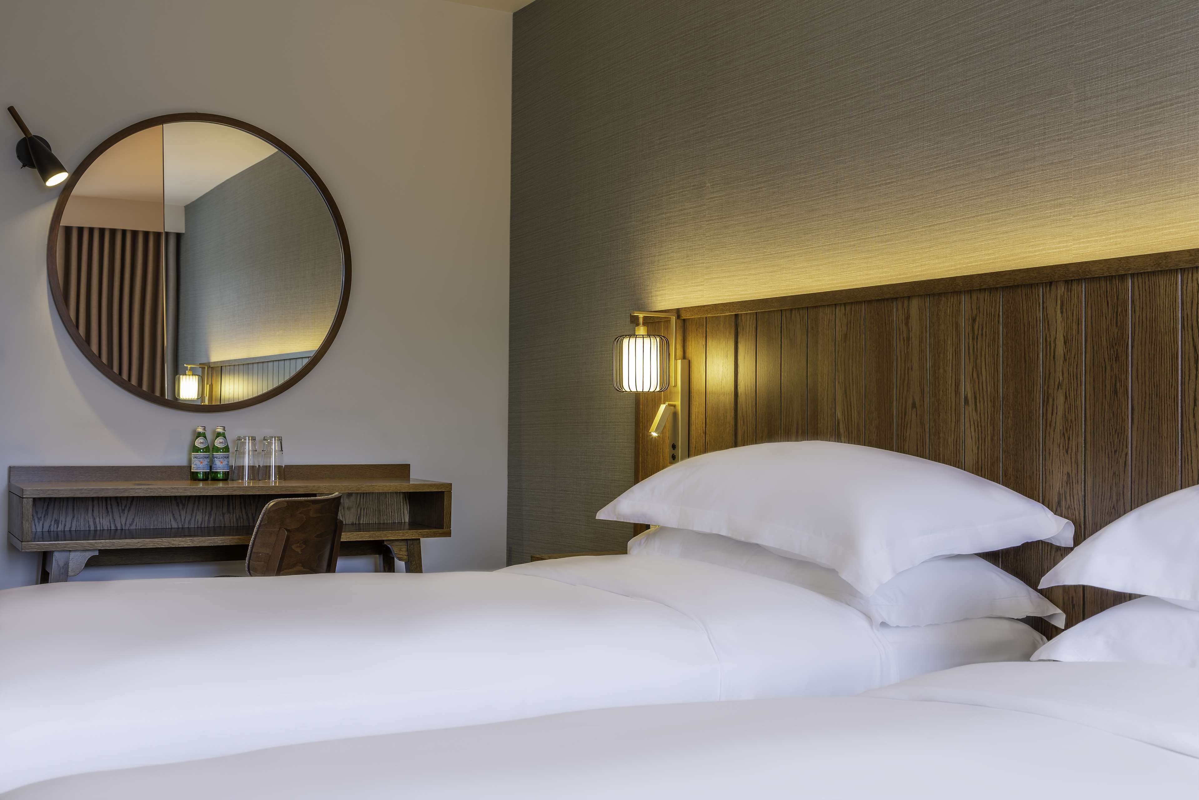 A hotel room with a large round mirror, a wooden headboard, and a bedside table with two bottles and glasses.