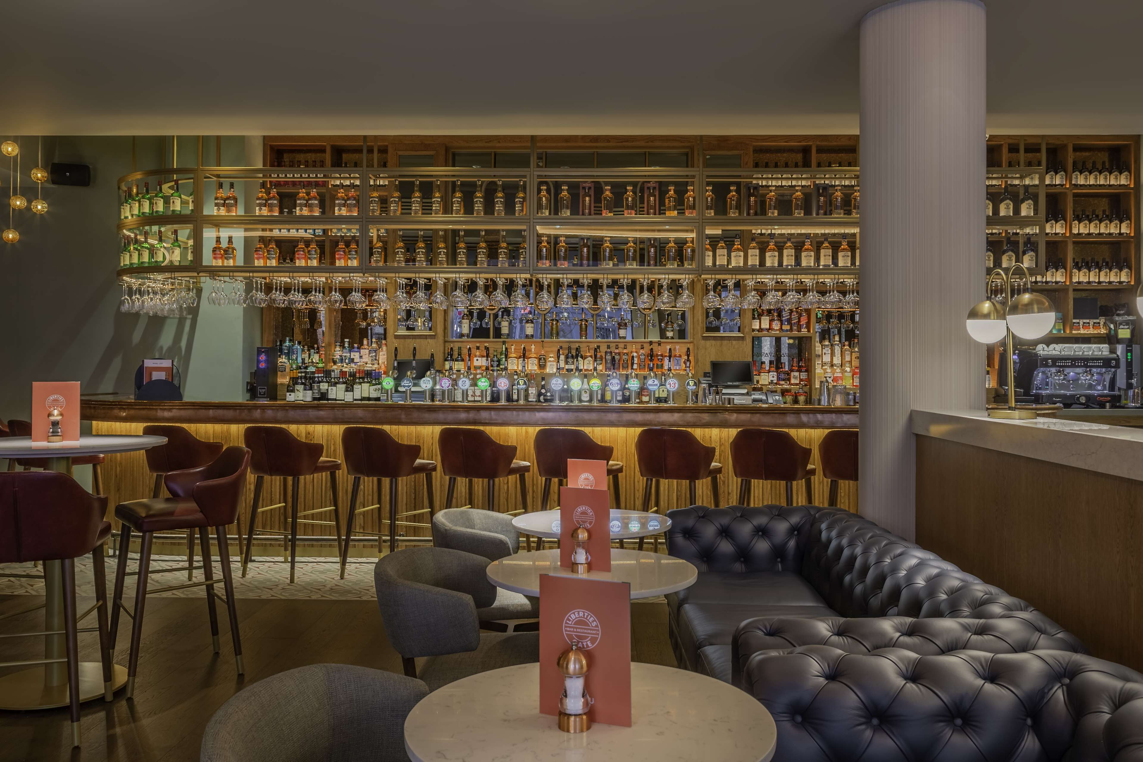 A bar area with multiple seating options including round tables and a sofa, a well-stocked bar with various bottles and glasses, and a large column in the foreground.