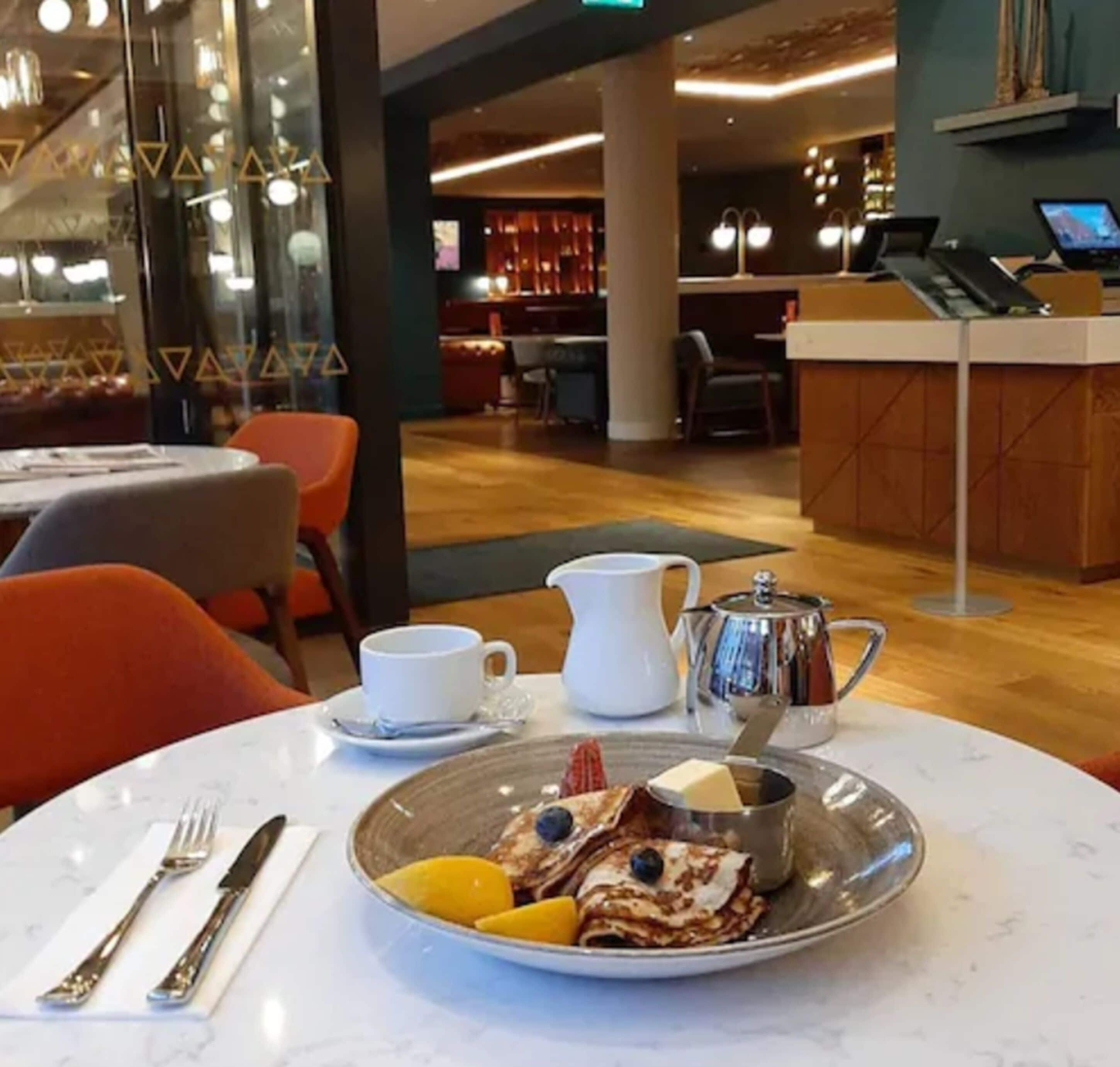 A dining area with a table set for breakfast, including a plate of crepes, a pitcher, a cup, and a spoon.