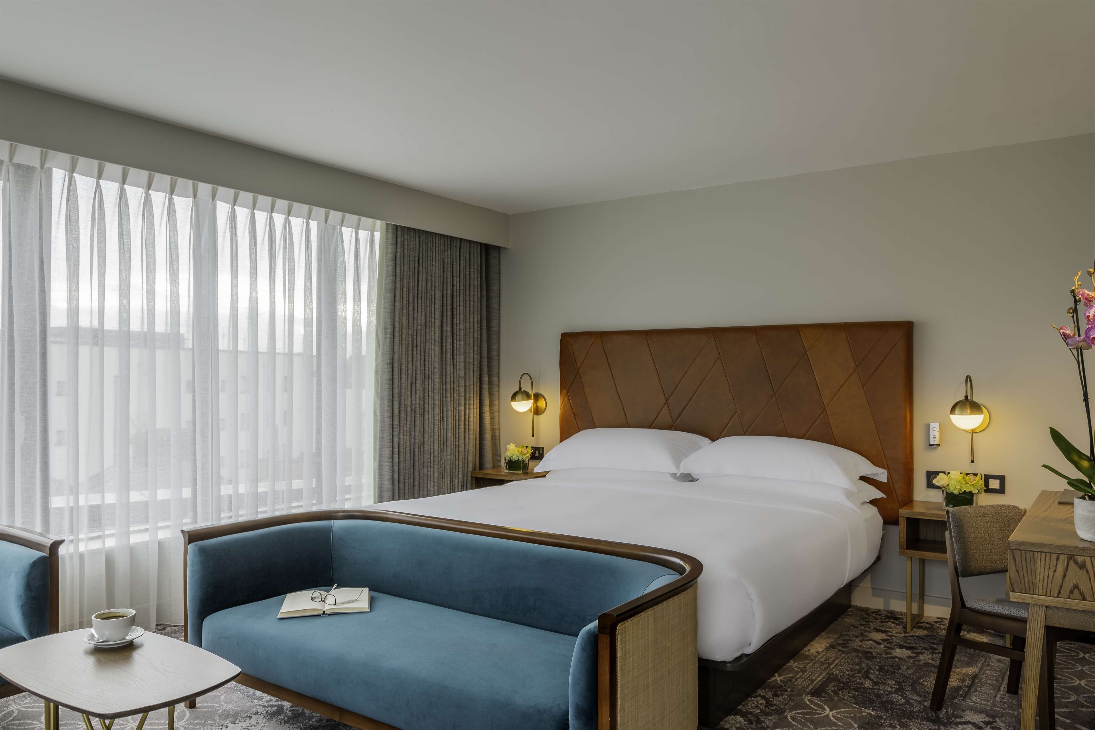 Een hotelkamer met een bed, een blauwe bank, een klein tafeltje met een kopje en een bureau met een lamp.