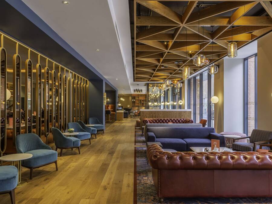 A modern hotel lobby with seating areas, a bar, and a kitchen in the background.