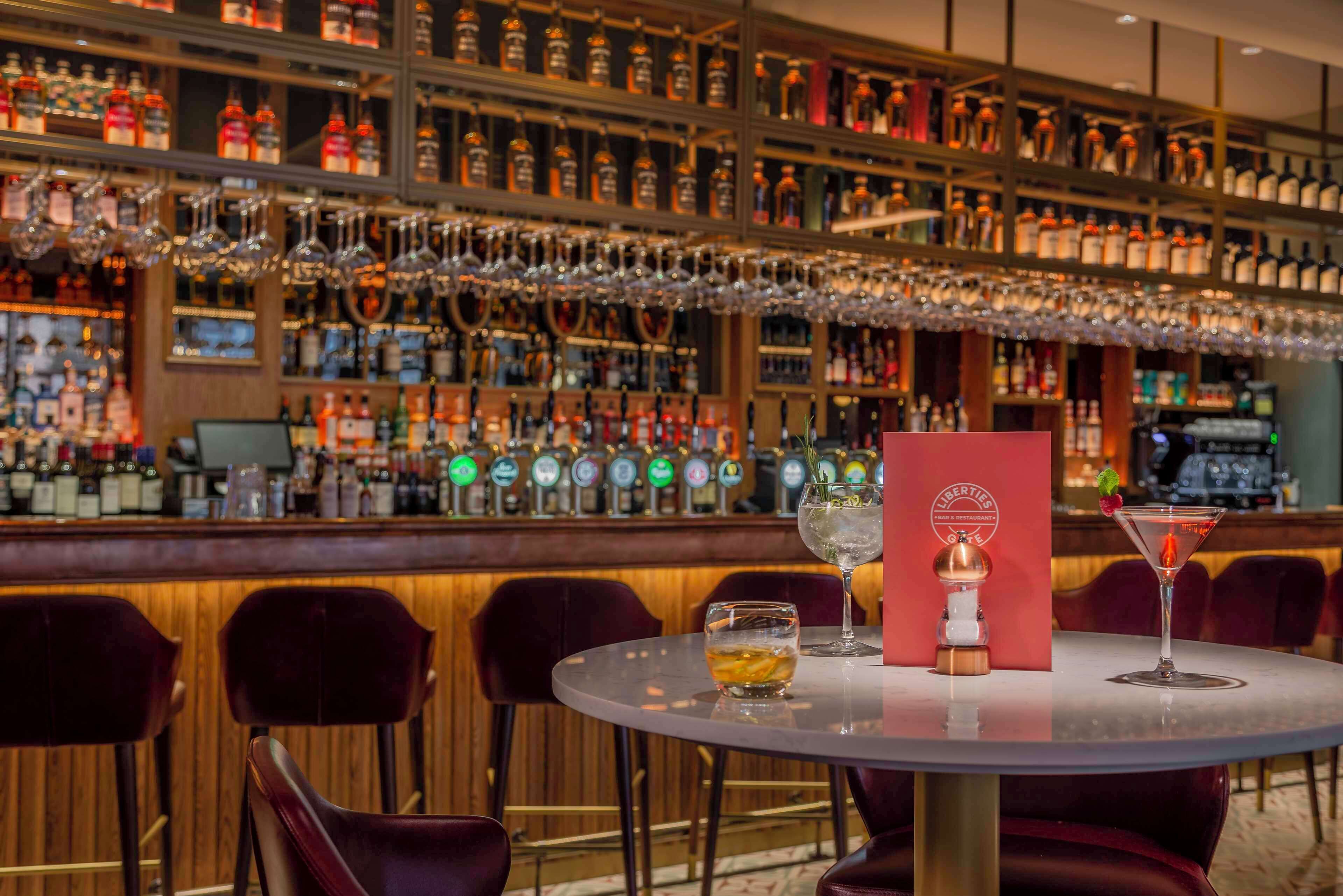 Zona de bar con varias botellas de licor y copas en estanterías, una barra y una mesa redonda con sillas.