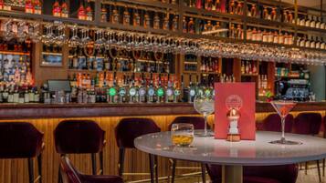 Zona de bar con varias botellas de licor y copas en estanterías, una barra y una mesa redonda con sillas.