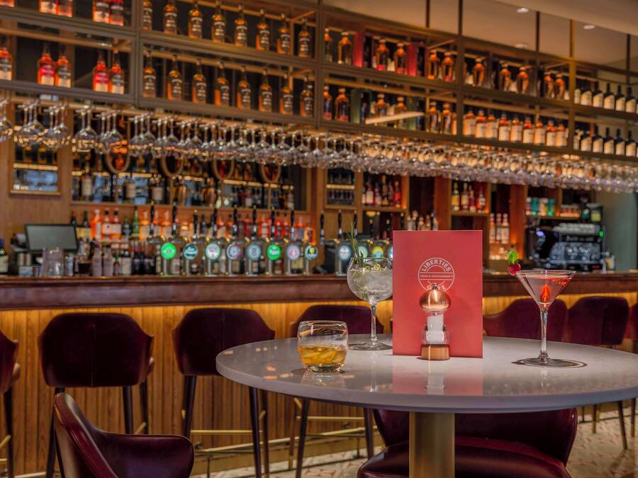 A bar area with multiple liquor bottles and glasses on shelves, a bar counter, and a round table with chairs.