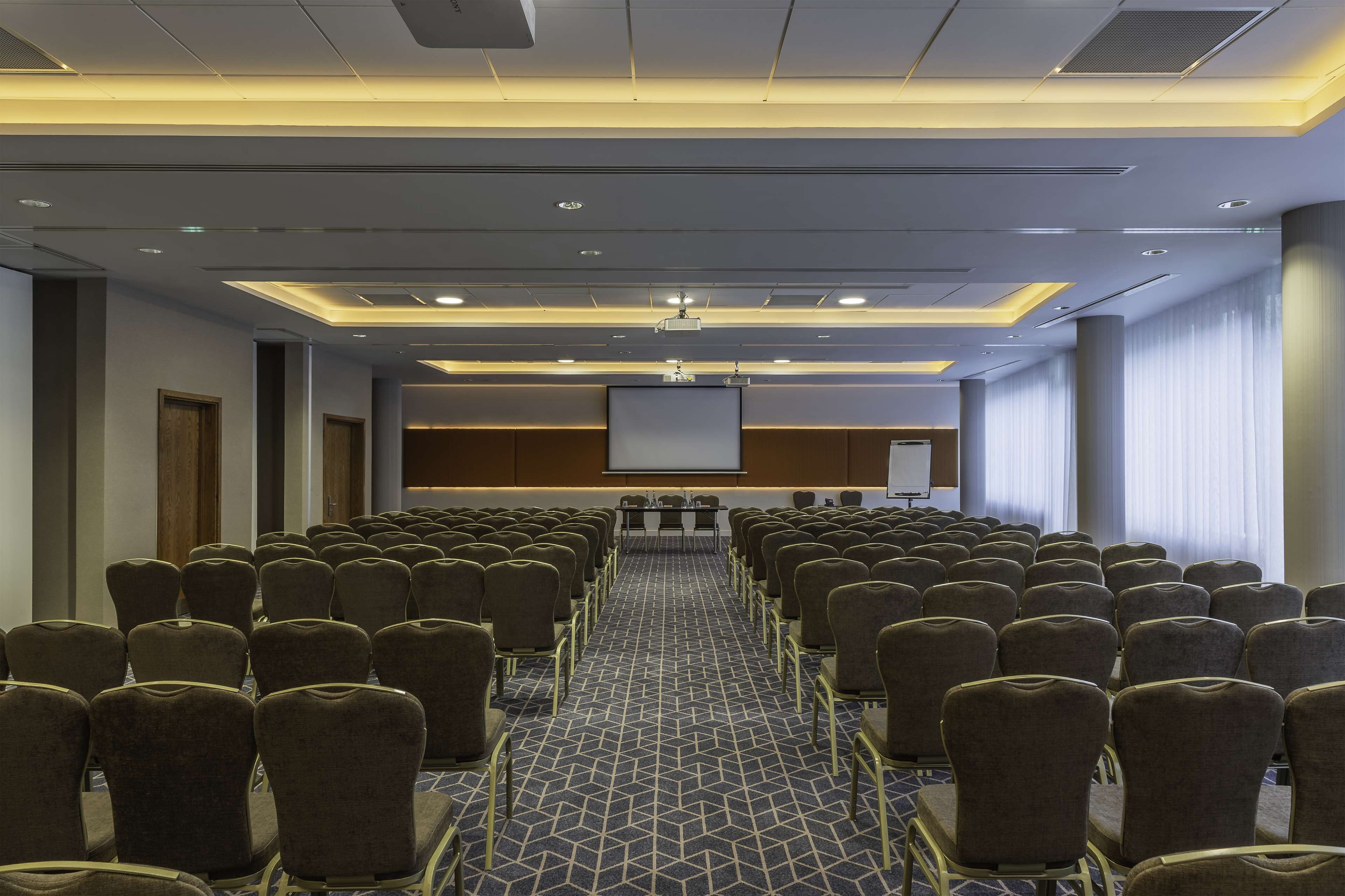 Una sala conferenze con file di sedie, uno schermo per proiezioni e un'area per presentazioni.