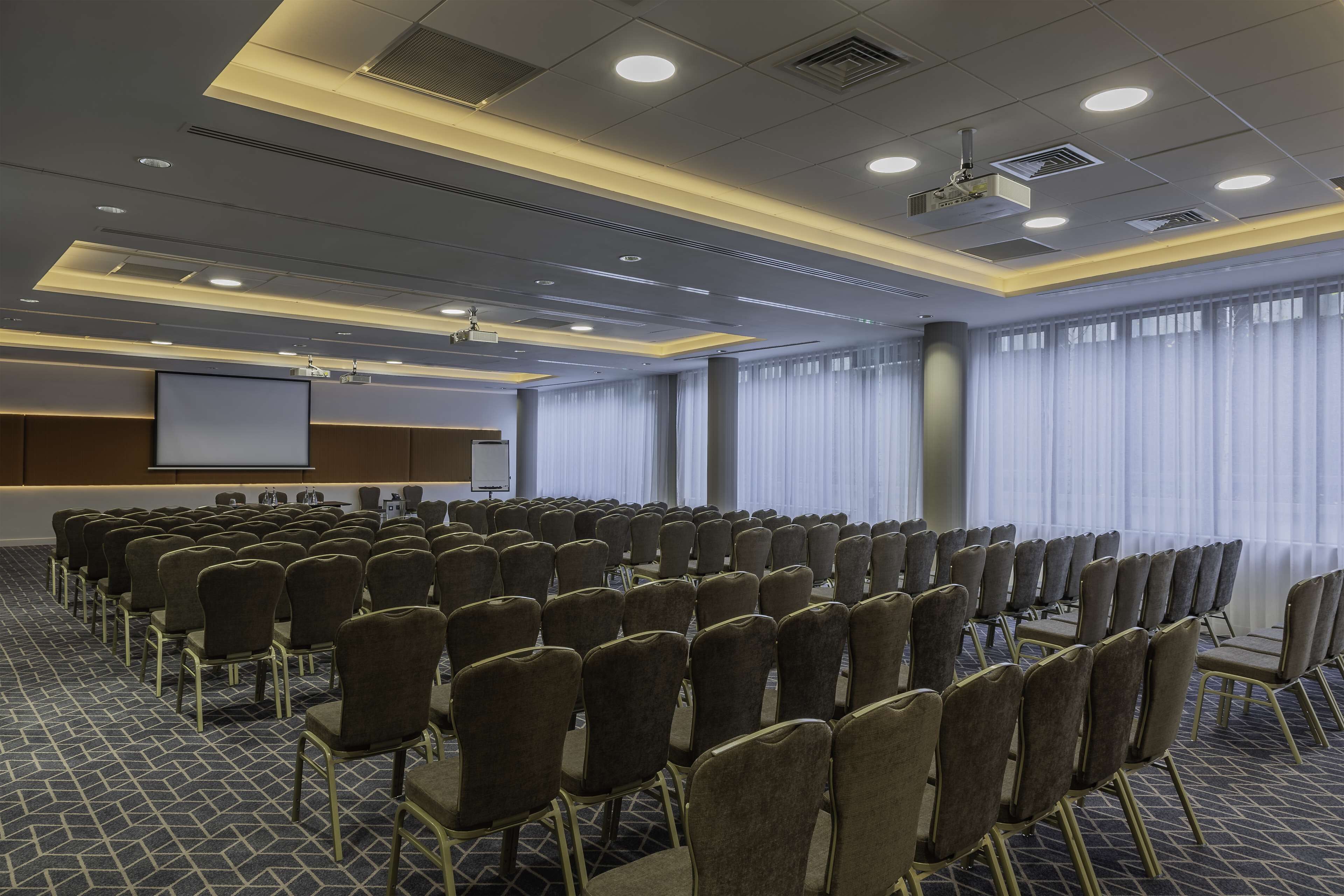 Un salón de conferencias con filas de sillas, una pantalla de proyector y un amplio ventanal con cortinas.