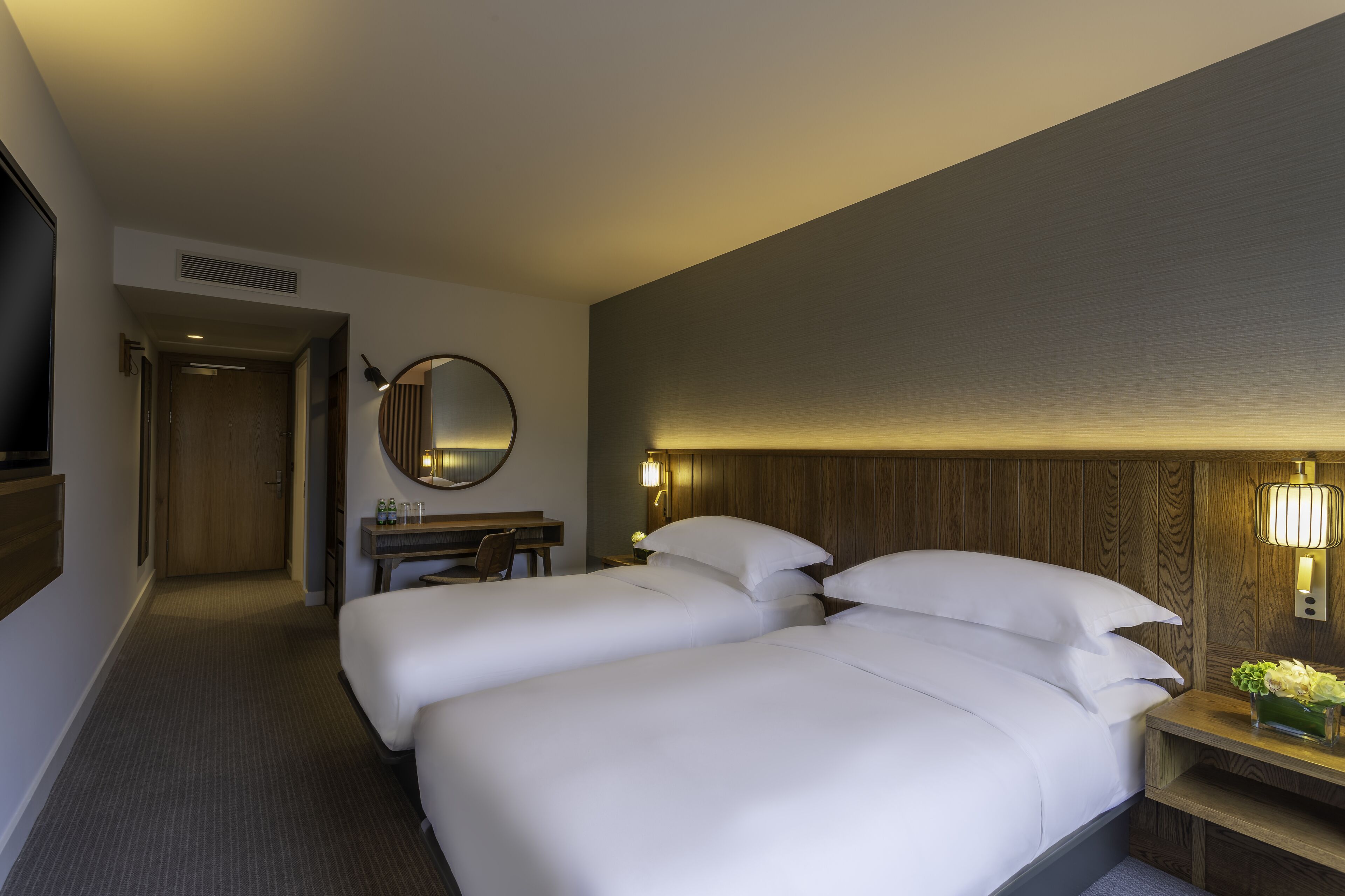 Quarto de hotel com duas camas, uma escrivaninha, um espelho e uma mesa de cabeceira com luminária.