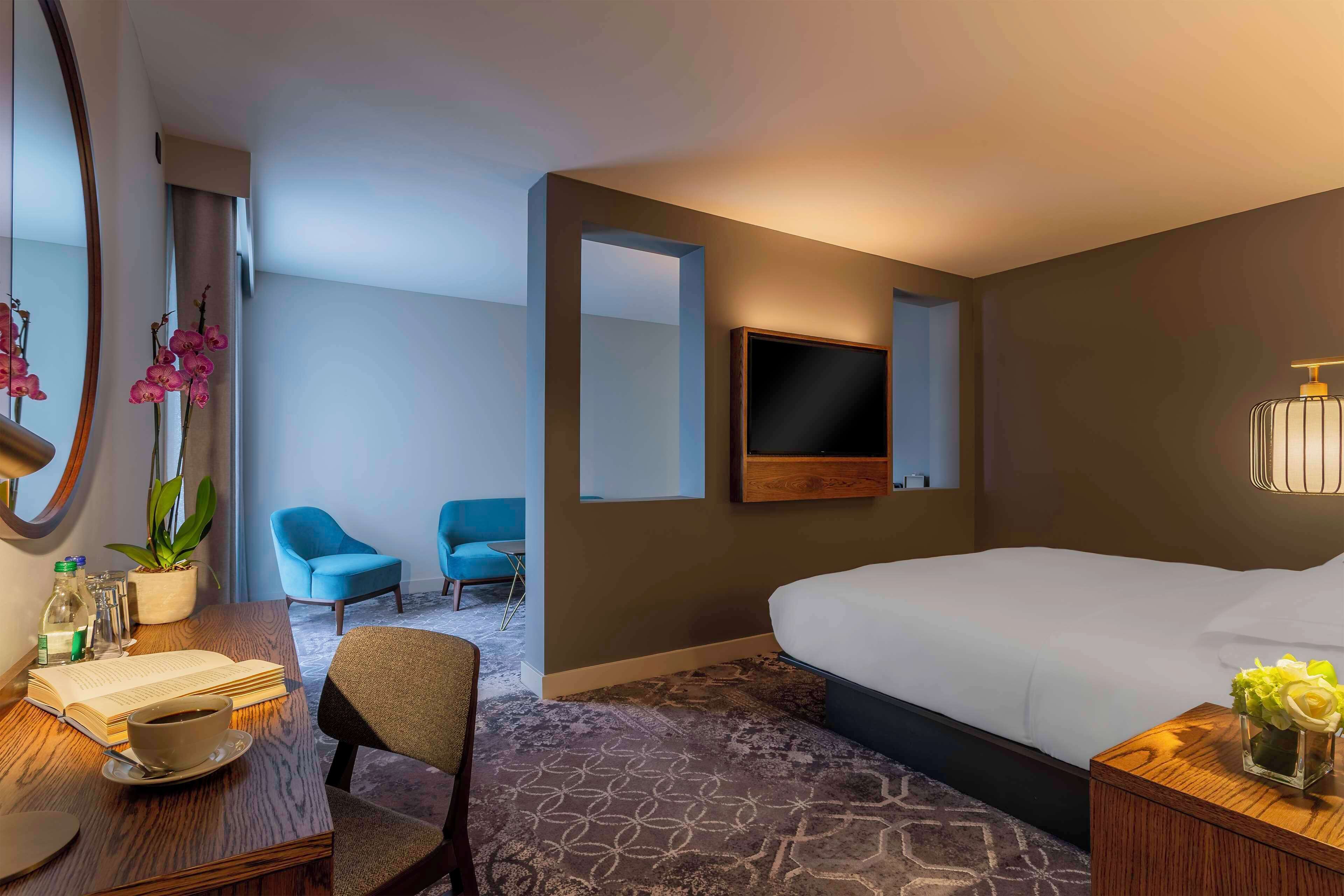 A hotel room with a large bed, a desk with a chair, a TV mounted on the wall, and a small table with a vase of flowers.