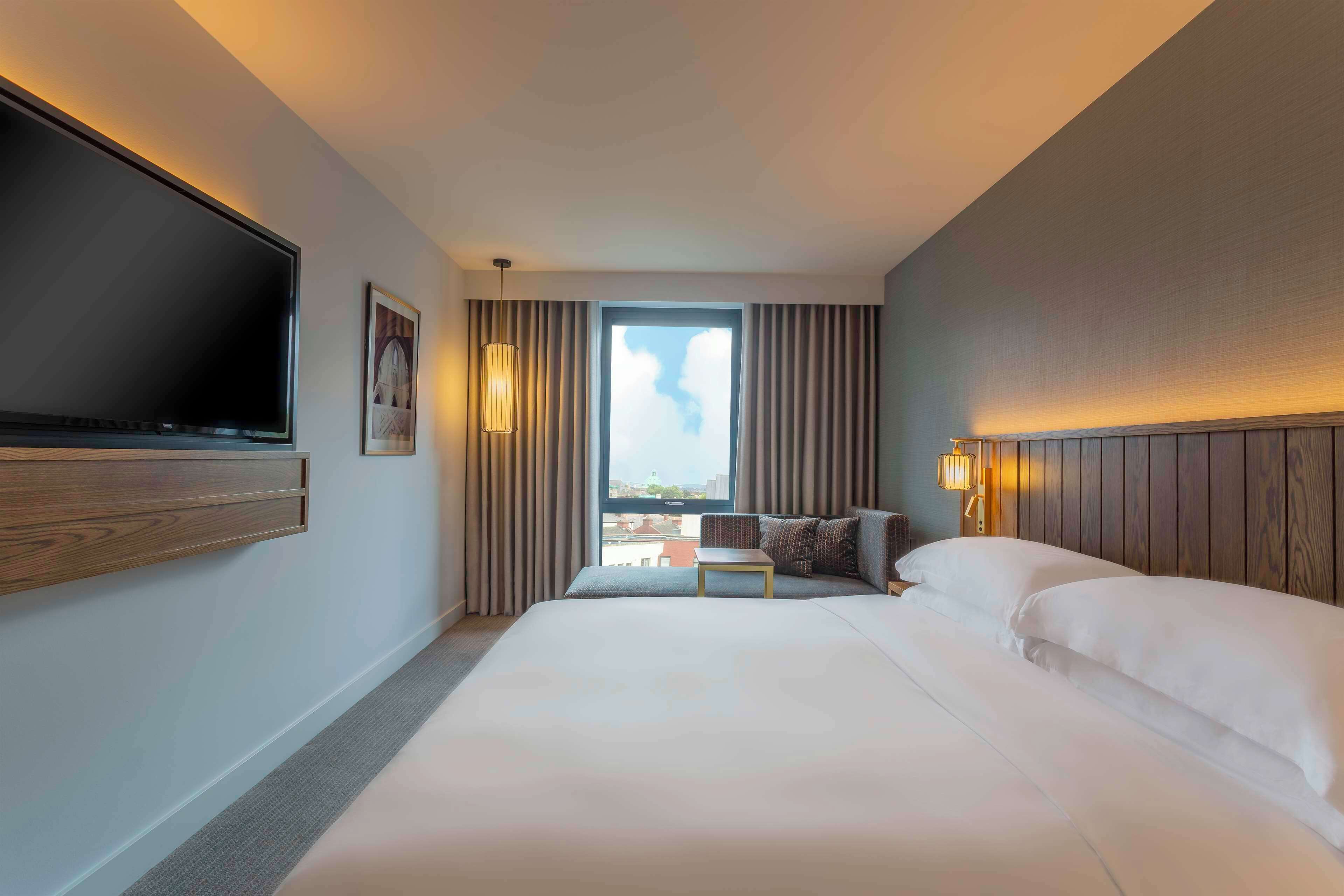 Habitación de hotel con una cama grande, un televisor de pantalla plana en la pared, una mesita y vistas a la ciudad desde la ventana.