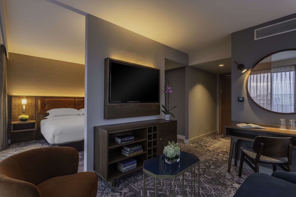 Een hotelkamer met een bed, een aan de muur bevestigde televisie, een bureau met stoel, een bijzettafeltje met vaas en een boekenkast.