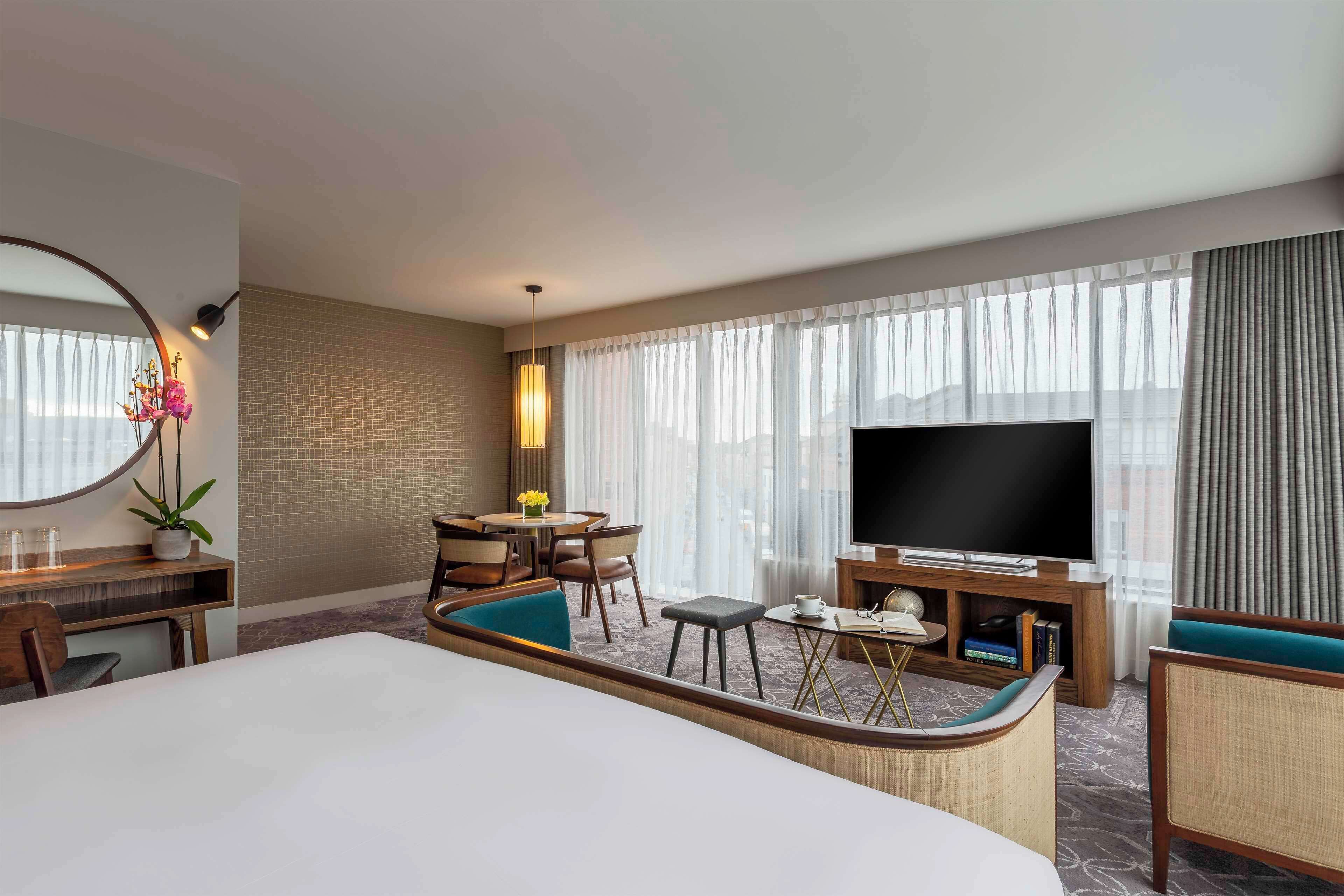 Quarto de hotel com uma cama grande, televisão, área de estar com mesa e cadeiras, e vista para a cidade pela janela.