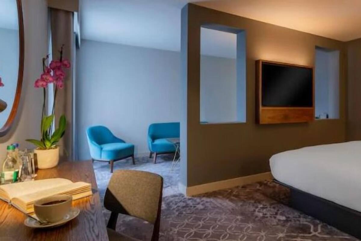 Een hotelkamer met een groot bed, een bureau met een stoel, een aan de muur bevestigde televisie en twee blauwe stoelen.