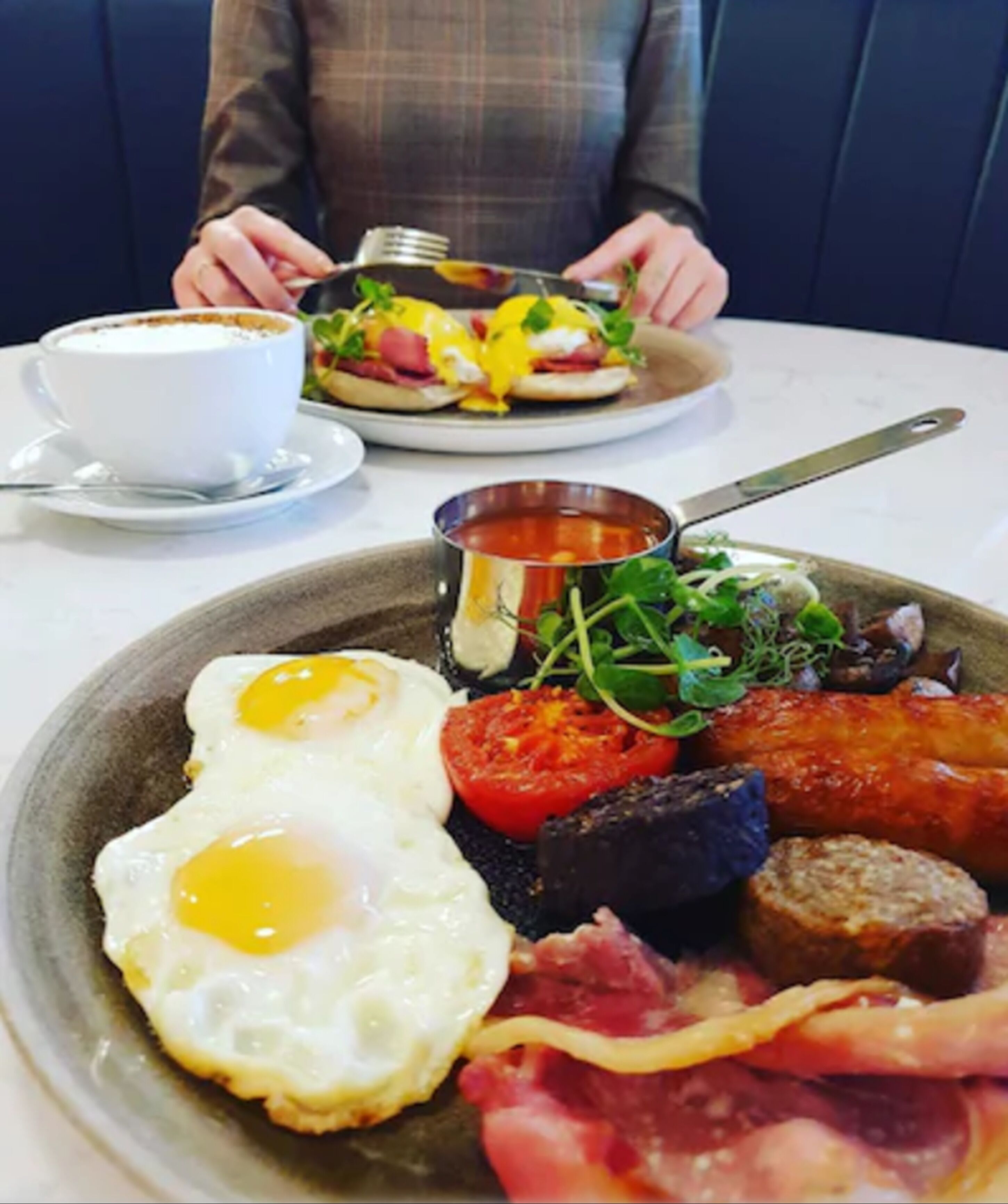 Ein Frühstücksteller mit Eiern, Speck, Würstchen, Tomaten und Salat, dazu eine Tasse Kaffee und eine kleine Schale Suppe.