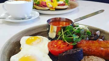 Ein Frühstücksteller mit Eiern, Speck, Würstchen, Tomaten und Salat, dazu eine Tasse Kaffee und eine kleine Schale Suppe.