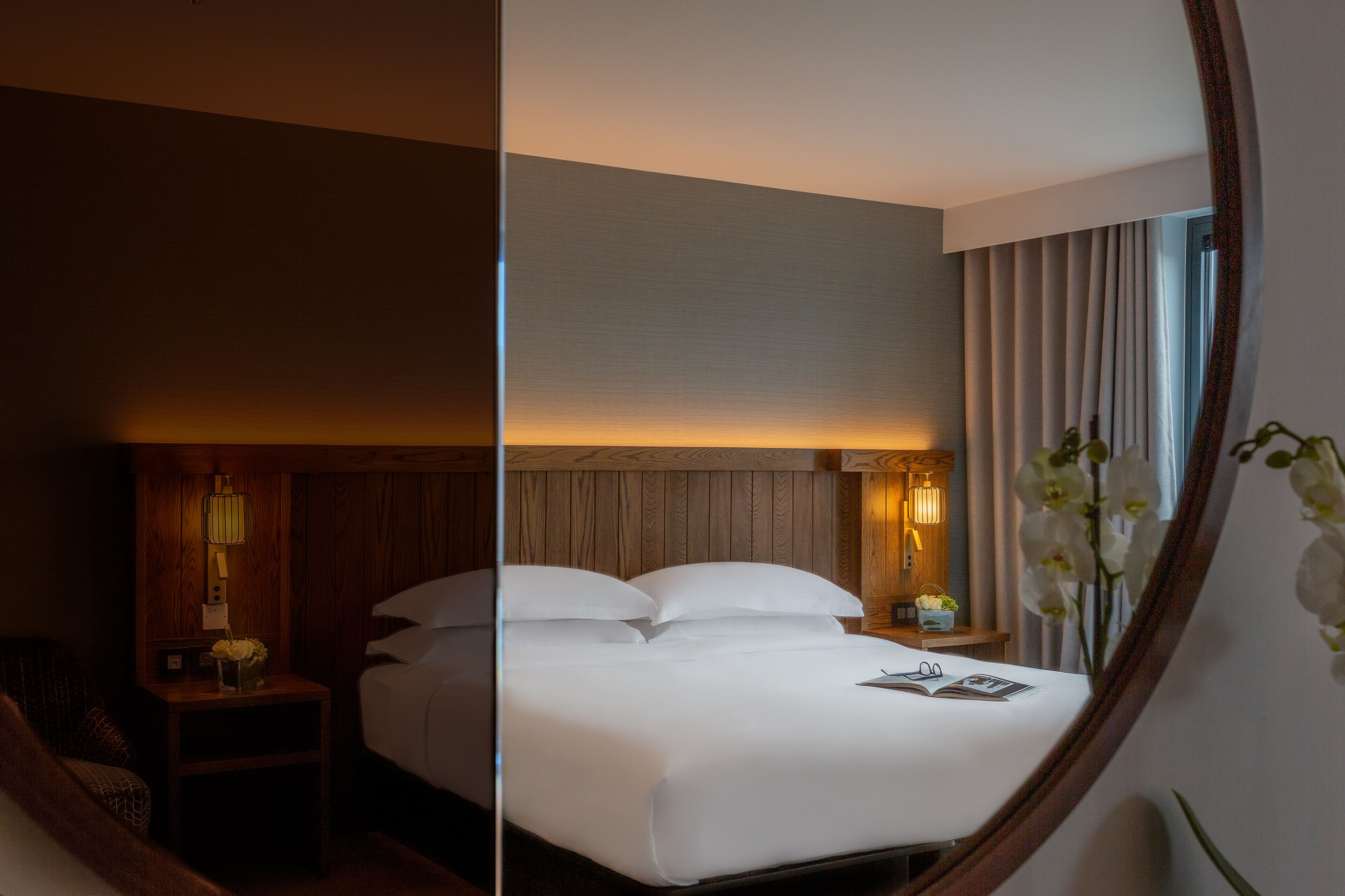 A hotel room with a large bed, a nightstand with a lamp, a small table with a vase of flowers, and a mirror reflecting the room.