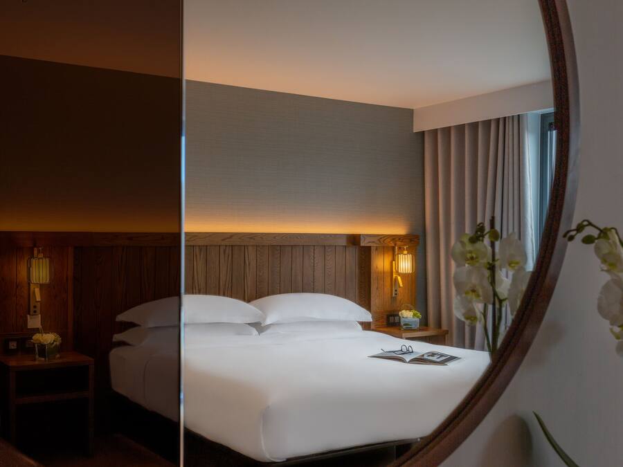 A hotel room with a large bed, a nightstand with a lamp, a small table with a vase of flowers, and a mirror reflecting the room.