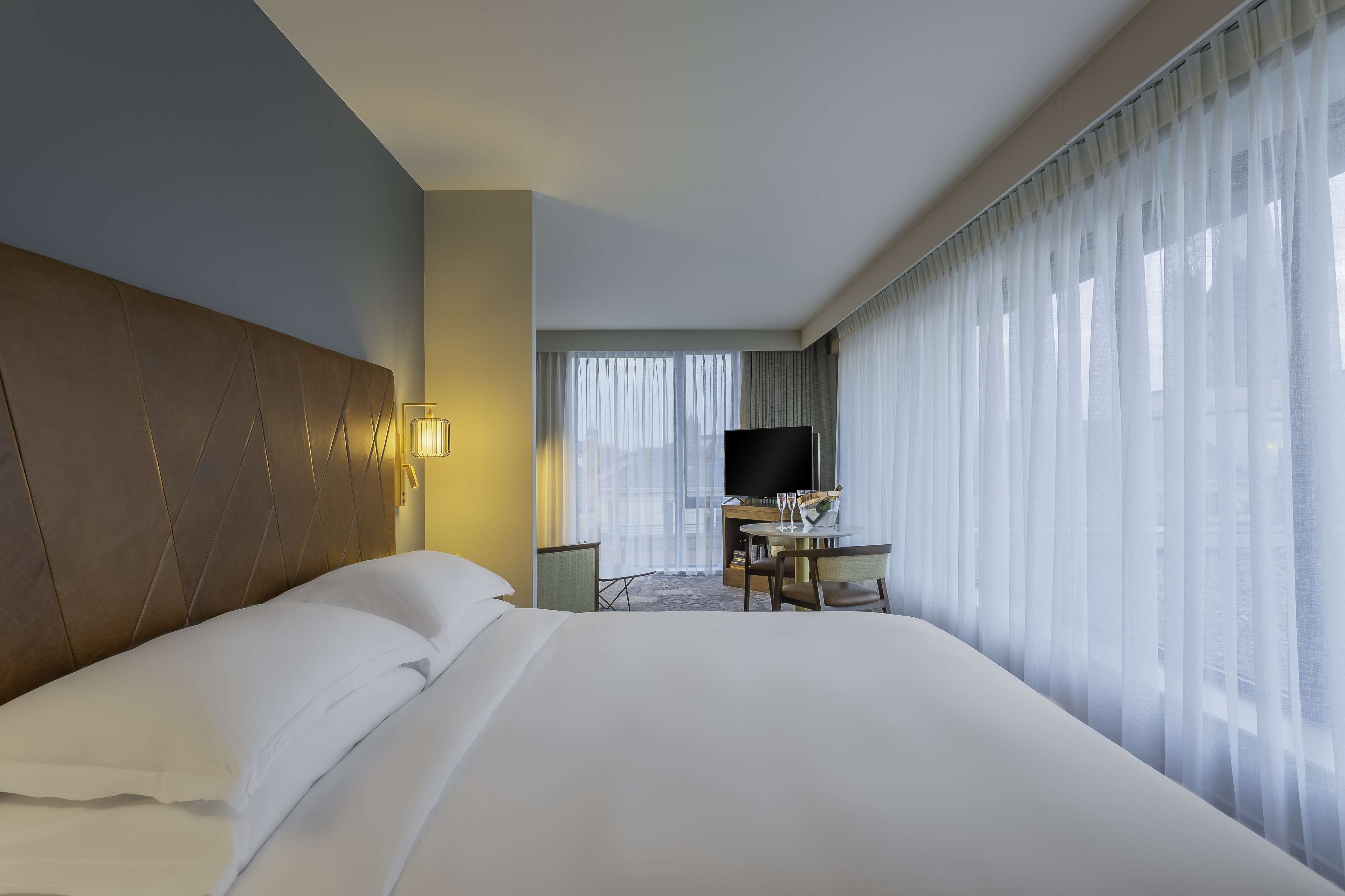 Una habitación de hotel con una cama grande, un escritorio con silla, un televisor y una ventana con cortinas translúcidas.
