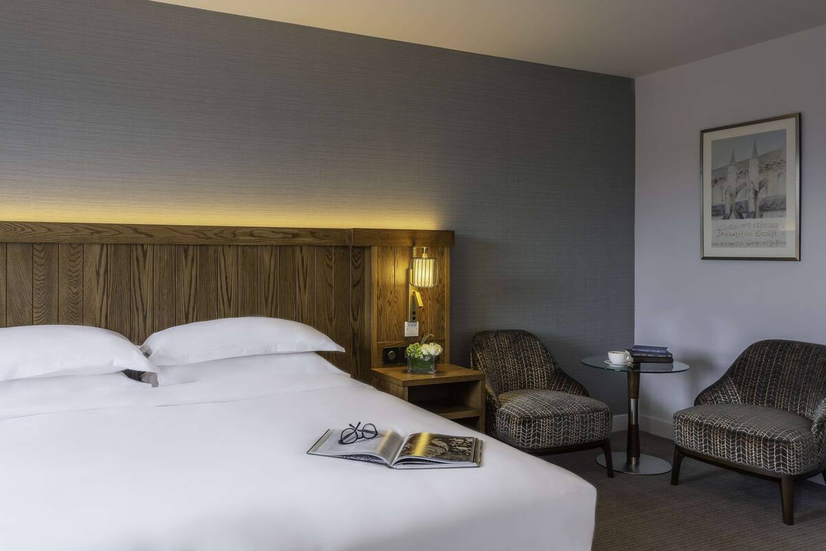 Een hotelkamer met een groot bed, twee stoelen, een klein tafeltje en een nachtkastje met een lamp en bloemen.