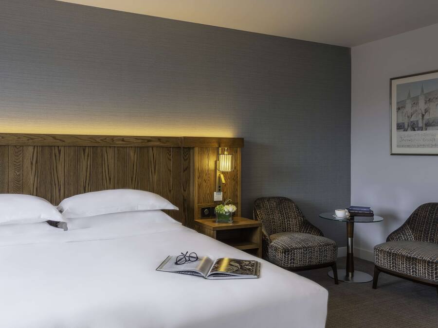 A hotel room with a large bed, two chairs, a small table, and a nightstand with a lamp and flowers.