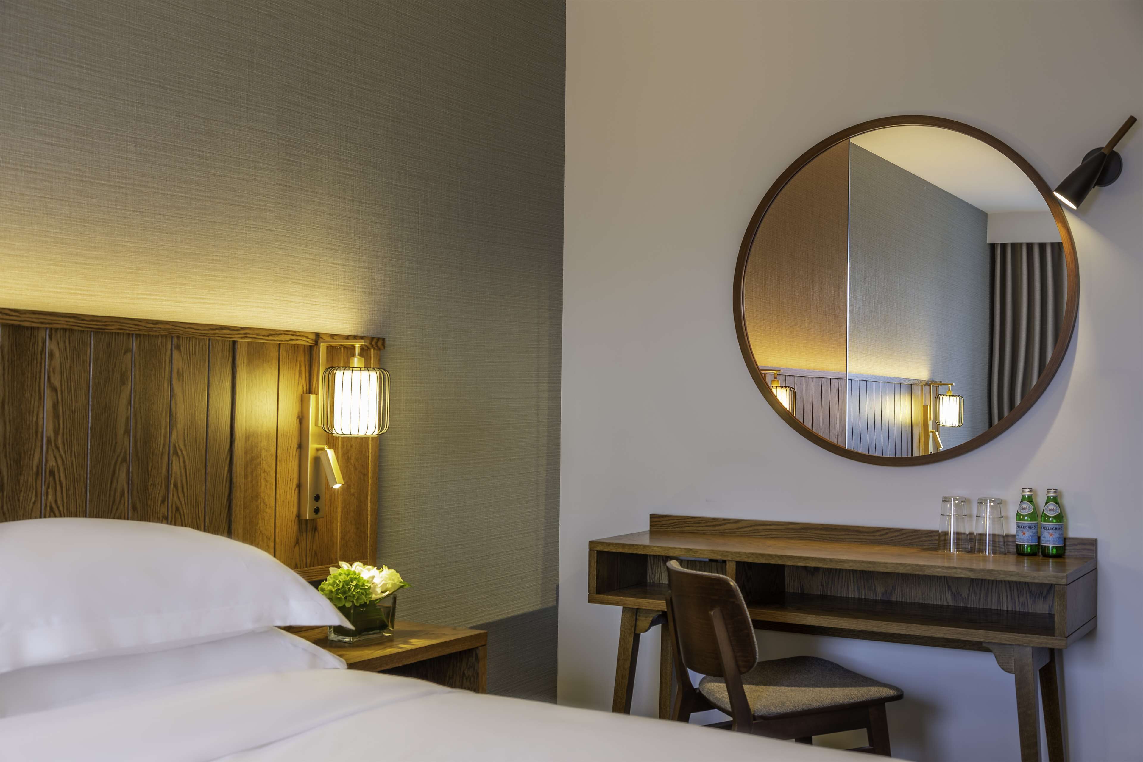 A hotel room with a bed, a desk, a chair, a round mirror, and a nightstand with a lamp.