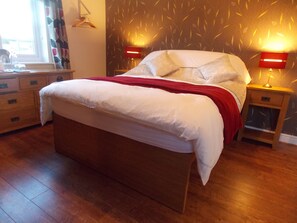 A neatly made bed with a red blanket, two bedside tables with lamps, a wooden dresser, and a patterned wall. - Craig Eithin B & B Holyhead (Holyhead)