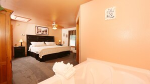 A bedroom with a bed, a nightstand, a bedside table, a ceiling fan, a window with curtains, and a bathroom with a bathtub and towels.