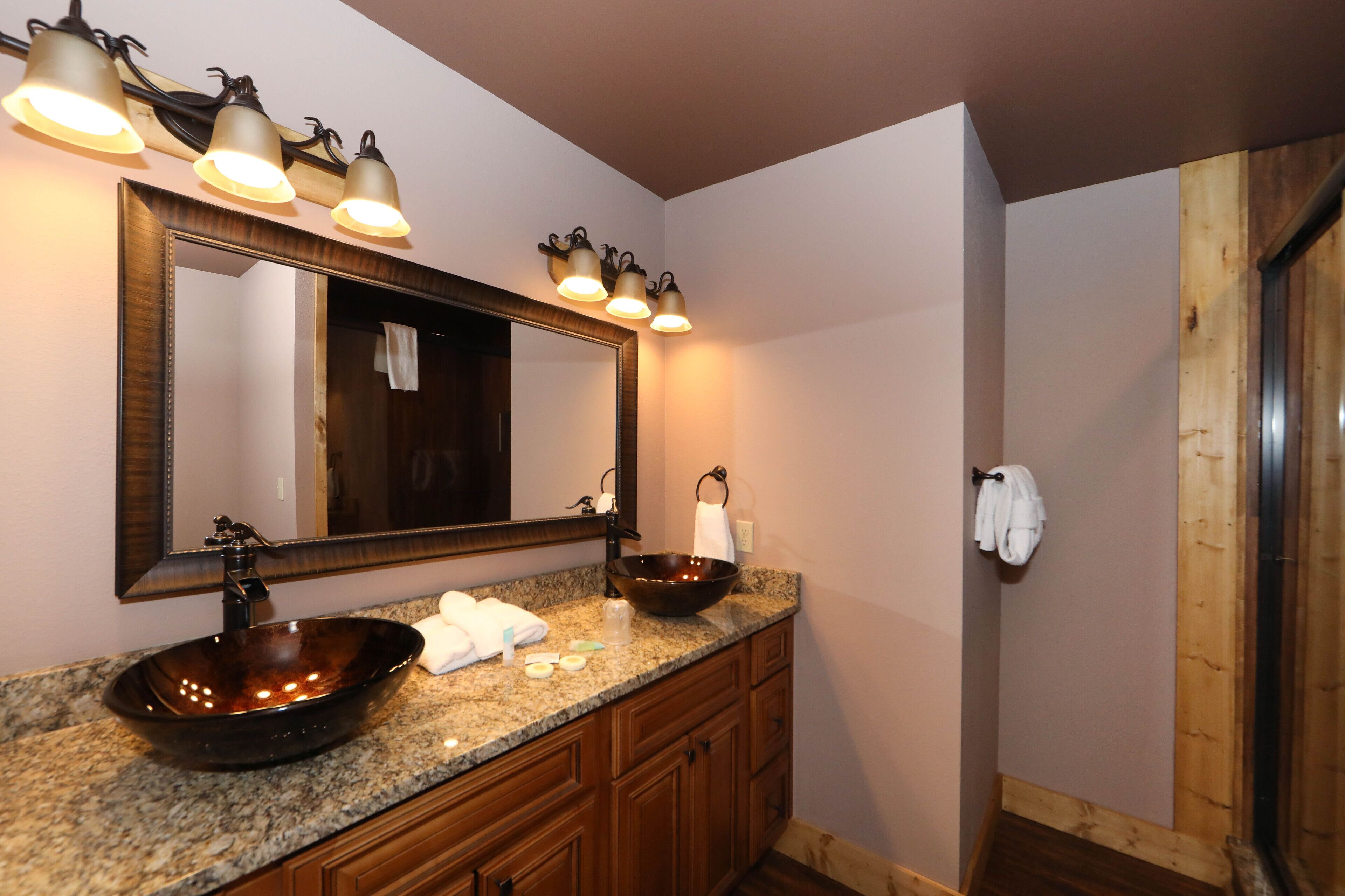 Une salle de bain dotée d’un comptoir en granit, d’un grand miroir, d’un lavabo sur colonne et d’armoires en bois.
