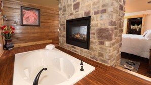 A bathroom with a large bathtub, a stone wall, a fireplace, and a wooden floor. - Above It All - Four Bedroom Cabin (Gatlinburg)