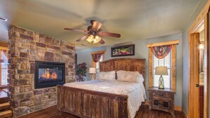 A bedroom with a stone fireplace, a wooden bed, a ceiling fan, and a nightstand with a lamp. - Aqua Paradise - Six Bedroom Chalet (Gatlinburg)