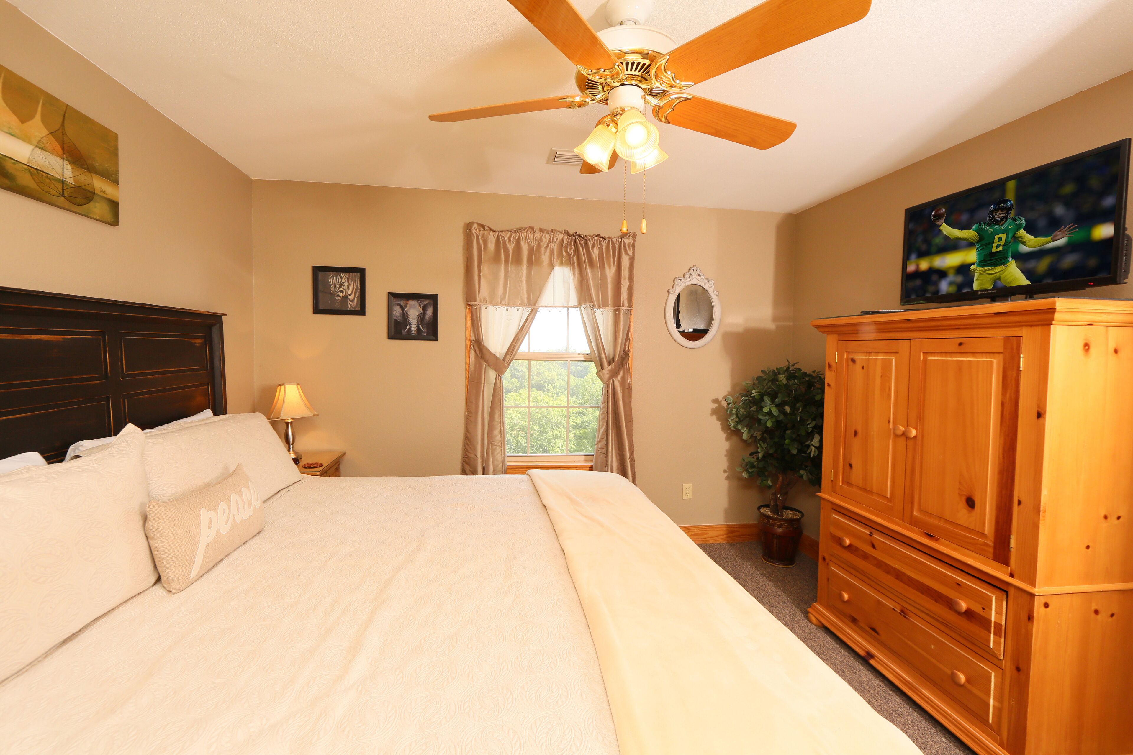 A bedroom with a large bed, a ceiling fan, a TV, a wooden dresser, and a window with curtains.