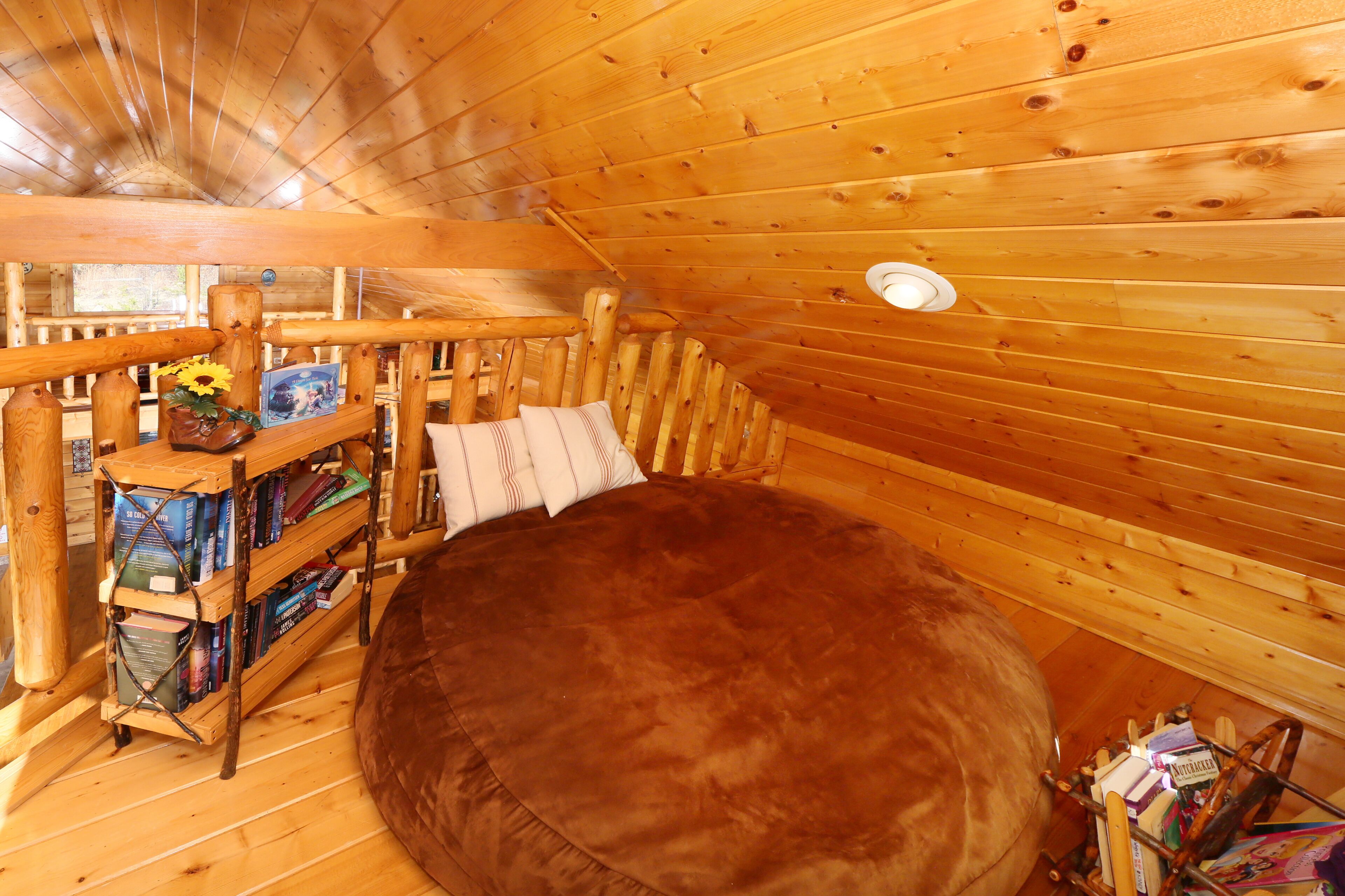 A cozy wooden cabin room with a round, plush ottoman, a wooden bookshelf, and a simple wooden railing.