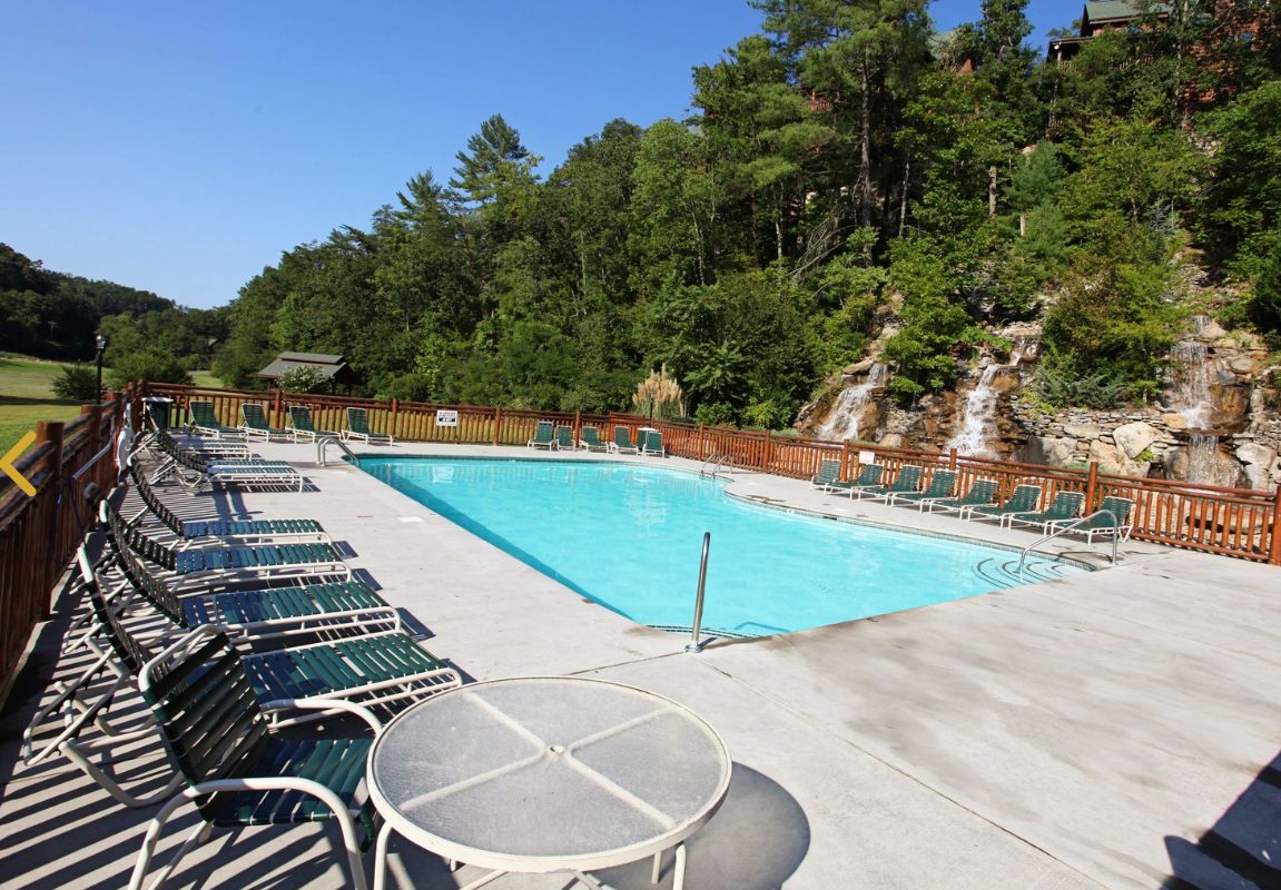 A rectangular swimming pool with a wooden deck, surrounded by lounge chairs and a table.