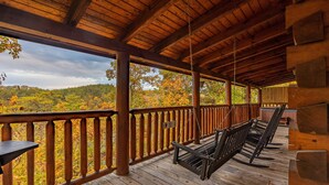 A wooden deck with a swing chair and a table, overlooking a forested area with autumn foliage.