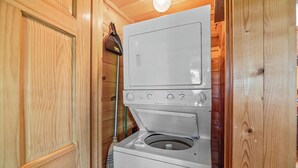 A compact laundry room with a stacked washer and dryer, wooden walls, and a broom leaning against the wall.