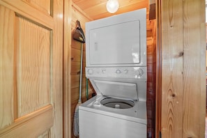 A compact laundry room with a stacked washer and dryer, wooden walls, and a broom leaning against the wall.