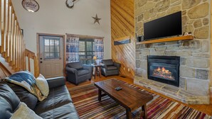 A living room with a stone fireplace, a television mounted above it, and a banjo on the wall. - Divine Escape - Two Bedroom Chalet (Gatlinburg)